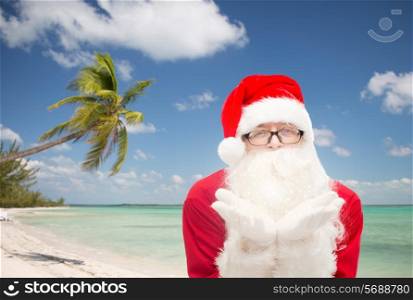 christmas, holidays, travel and people concept - man in costume of santa claus blowing on palms over tropical beach background