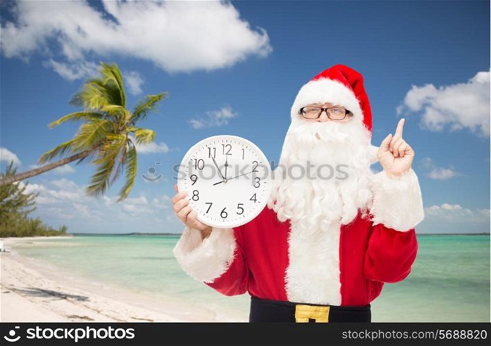 christmas, holidays, travel and people concept - man in costume of santa claus with clock showing twelve pointing finger up over tropical beach background