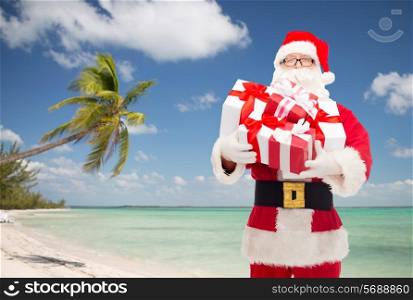 christmas, holidays, travel and people concept - man in costume of santa claus with gift boxes over tropical beach background