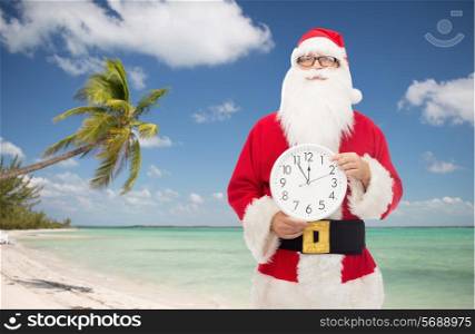 christmas, holidays, travel and people concept - man in costume of santa claus with clock showing twelve pointing finger over tropical beach background