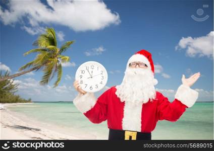 christmas, holidays, travel and people concept - man in costume of santa claus with clock showing twelve over tropical beach background