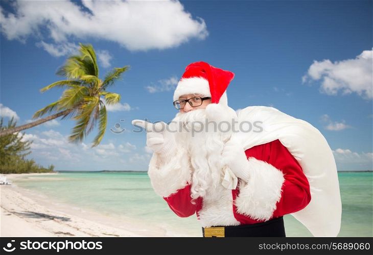 christmas, holidays, travel and people concept - man in costume of santa claus with bag pointing finger over tropical beach background