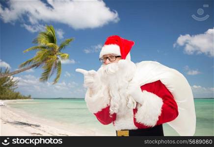 christmas, holidays, travel and people concept - man in costume of santa claus with bag pointing finger over tropical beach background