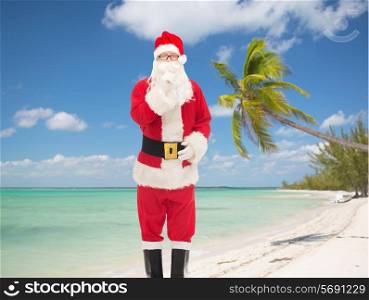 christmas, holidays, travel and people concept - man in costume of santa claus making hush gesture over tropical beach background