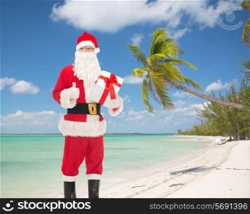 christmas, holidays, travel and people concept - man in costume of santa claus with gift box showing thumbs up gesture over tropical beach background