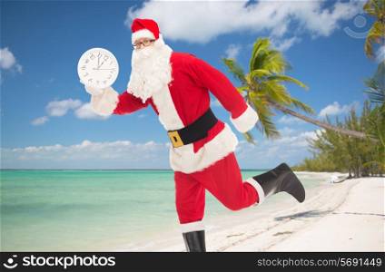 christmas, holidays, travel and people concept - man in costume of santa claus running with clock showing twelve over tropical beach background