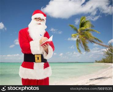christmas, holidays, travel and people concept - man in costume of santa claus with notepad and pen over tropical beach background