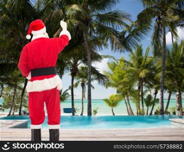 christmas, holidays, travel and people concept - man in costume of santa claus writing something from back over tropical beach and swimming pool background