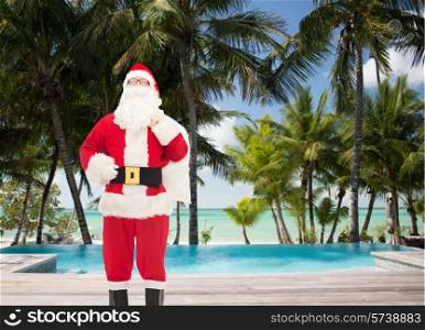christmas, holidays, travel and people concept - man in costume of santa claus with bag over tropical beach and swimming pool background