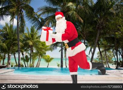 christmas, holidays, travel and people concept - man in costume of santa claus running with gift box over tropical beach and swimming pool background