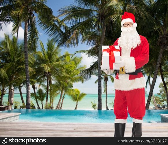 christmas, holidays, travel and people concept - man in costume of santa claus with gift box over tropical beach and swimming pool background