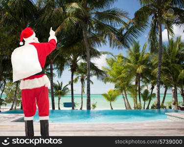 christmas, holidays, travel and people concept - man in costume of santa claus with bag pointing finger from back over tropical beach and swimming pool background
