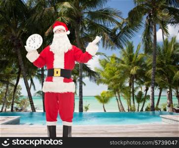 christmas, holidays, travel and people concept - man in costume of santa claus with clock showing twelve over tropical beach and swimming pool background