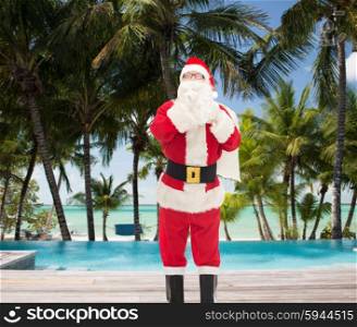christmas, holidays, travel and people concept - man in costume of santa claus with bag making hush gesture over swimming pool on tropical beach background
