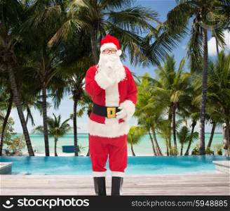 christmas, holidays, travel and people concept - man in costume of santa claus making hush gesture over swimming pool on tropical beach background