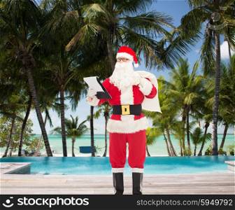 christmas, holidays, travel and people concept - man in costume of santa claus with notepad and bag over swimming pool on tropical beach background