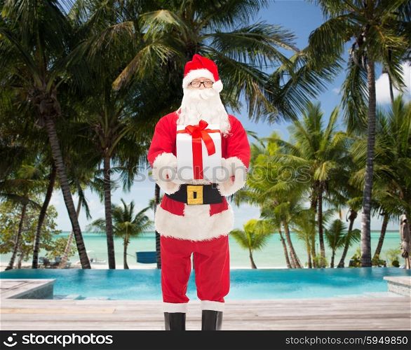 christmas, holidays, travel and people concept - man in costume of santa claus with gift box over swimming pool on tropical beach background