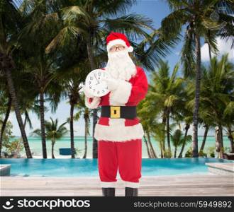 christmas, holidays, travel and people concept - man in costume of santa claus with clock showing twelve pointing finger over swimming pool on tropical beach background