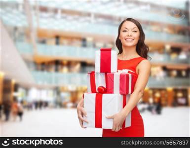 christmas, holidays, valentine&#39;s day, celebration and people concept - smiling woman in red dress with gift boxes over shopping center background