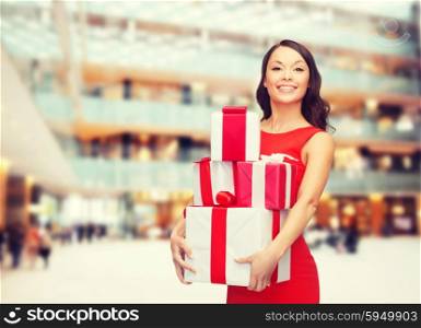 christmas, holidays, valentine&amp;#39;s day, celebration and people concept - smiling woman in red dress with gift boxes over shopping center background