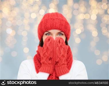 christmas, holidays, winter and people concept - surprised woman in hat, muffler and mittens over lights background