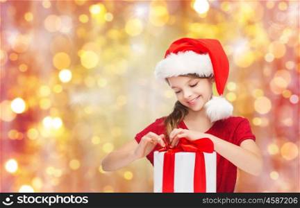 christmas, holidays, winter, children and people concept - smiling girl in santa helper hat with gift box over lights background