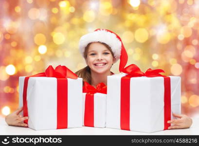 christmas, holidays, winter, children and people concept - smiling girl in santa helper hat with gift boxes over lights background