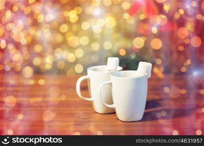 christmas, holidays, winter, food and drinks concept - close up of cups with hot chocolate or cocoa drinks and marshmallow on wooden table over lights