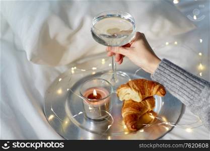 christmas, hygge and people concept - hand of woman holding champagne glass with croissants, candle and garland lights string in bed. hand of woman holding champagne with croissants