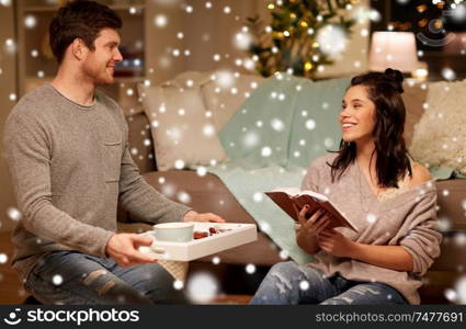 christmas, hygge and people concept - happy couple with book and food on tray at home over snow. happy couple with book and food at home