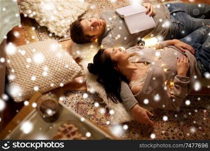 christmas, hygge and people concept - happy couple with book and garland lying on floor at home and sleeping over snow. happy couple with garland lying on floor at home