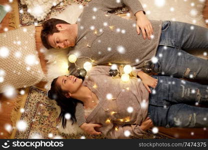 christmas, hygge and people concept - happy couple with garland lying on floor at home over snow. happy couple with garland lying on floor at home
