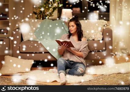 christmas, hygge and people concept - happy young woman reading book at home over snow. happy young woman reading book at home