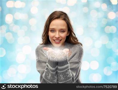 christmas, magic and holidays concept - happy young woman in grey ugly sweater holding fairy dust over festive lights on blue background. young woman in christmas sweater with fairy dust