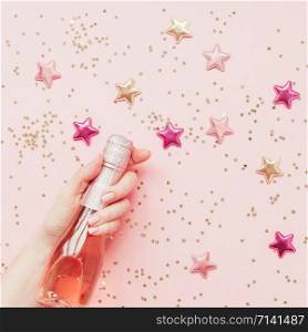 Christmas or New Year composition with bottle of rose champagne in woman hand and golden shiny sparkle star confetti on pastel pink background, top view. Celebration flat lay. Party creative concept