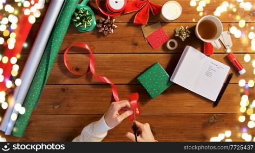 christmas, packing and holidays concept - hands with scissors cutting red ribbon for gift box on wooden table over festive lights. hands packing christmas gift and cutting ribbon