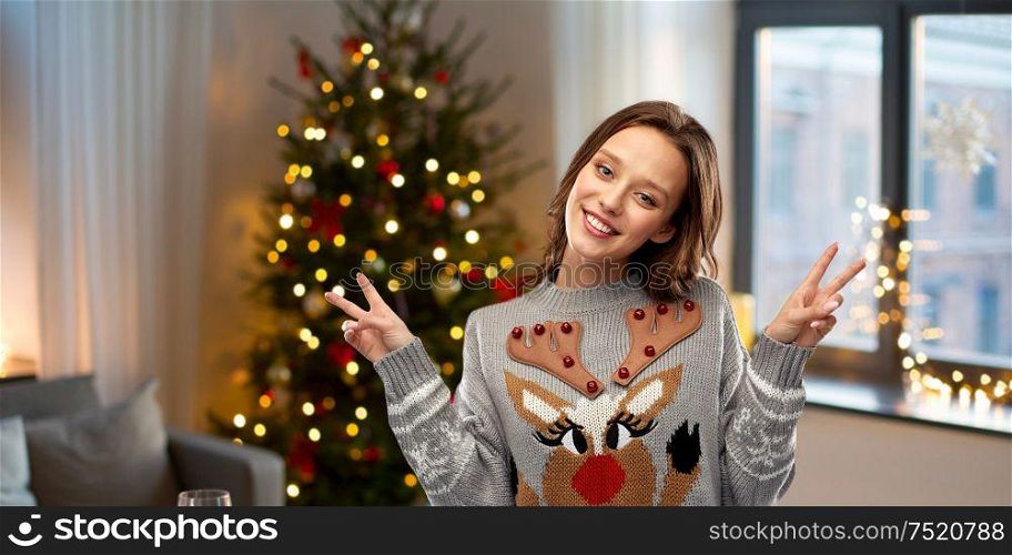 christmas, party and holidays concept - happy young woman wearing ugly sweater with reindeer pattern showing peace over home background. woman in ugly christmas sweater showing peace sign