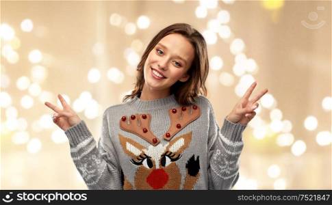 christmas, party and holidays concept - happy young woman wearing ugly sweater with reindeer pattern showing peace over grey background over festive lights background. woman in ugly christmas sweater showing peace sign