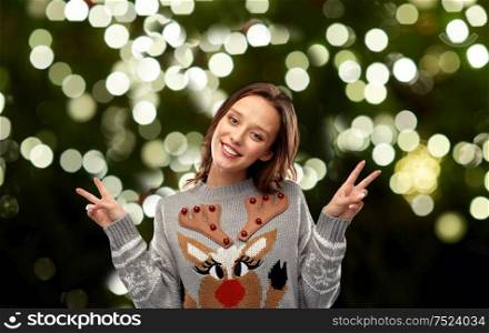christmas, party and holidays concept - happy young woman wearing ugly sweater with reindeer pattern showing peace over festive lights on dark green background. woman in ugly christmas sweater showing peace sign