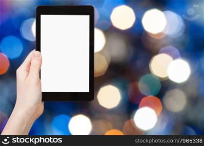 christmas party concept - hand with tablet pc with cut out screen on background from blue shimmering Christmas lights bokeh of electric garlands on Xmas tree