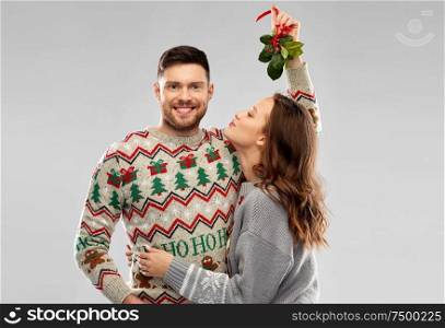 christmas, people and holiday traditions concept - portrait of happy couple in ugly sweaters kissing under the mistletoe over grey background. happy couple kissing under the mistletoe