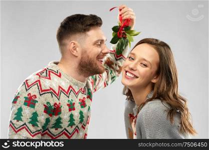 christmas, people and holiday traditions concept - portrait of happy couple in ugly sweaters kissing under the mistletoe over grey background. happy couple kissing under the mistletoe