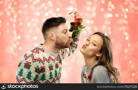 christmas, people and holiday traditions concept - portrait of happy couple in ugly sweaters kissing under mistletoe over festive lights on pink coral background. happy couple kissing under mistletoe on christmas