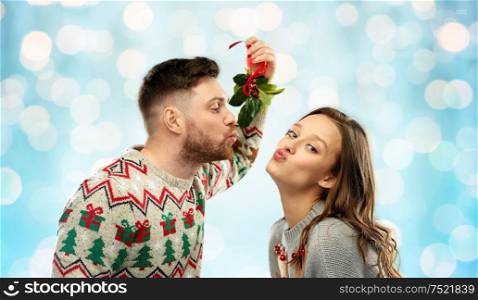 christmas, people and holiday traditions concept - portrait of happy couple in ugly sweaters kissing under mistletoe over festive lights on blue background. happy couple kissing under mistletoe on christmas