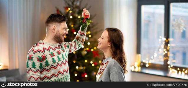christmas, people and holiday traditions concept- portrait of happy couple in ugly sweaters with mistletoe over home background. happy couple with mistletoe on christmas at home