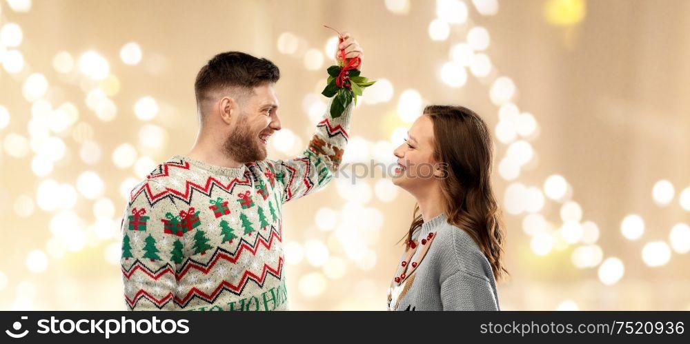 christmas, people and holiday traditions concept- portrait of happy couple in ugly sweaters with mistletoe over festive lights background. happy couple with mistletoe on christmas