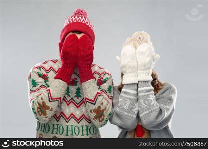 christmas, people and holidays concept - couple in ugly sweaters, hats and woolen mittens covering their faces over grey background. couple in ugly sweaters and mittens on christmas