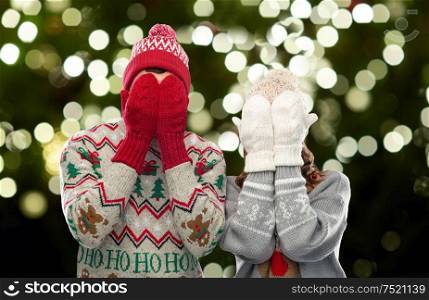 christmas, people and holidays concept - couple in ugly sweaters, hats and woolen mittens covering their faces over festive lights on dark green background. couple in ugly sweaters and mittens on christmas