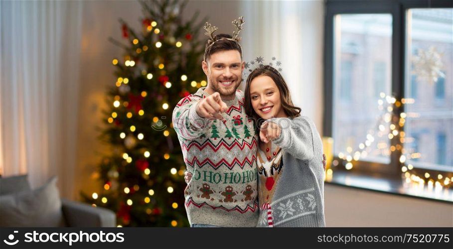 christmas, people and holidays concept - happy couple at ugly sweater party pointing at you over home background. happy couple at christmas ugly sweater party