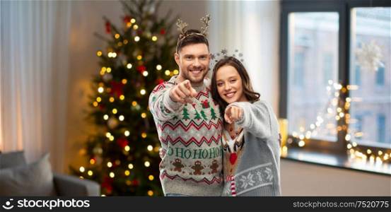 christmas, people and holidays concept - happy couple at ugly sweater party pointing at you over home background. happy couple at christmas ugly sweater party